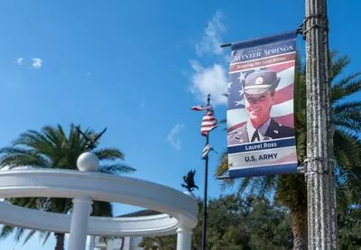 A vertical street banner mounted on a light pole reads “City of Winter Springs – Honoring Our Local Heroes” and features a portrait of Laurel Ross in a U.S. Army uniform against an American flag background. Palm trees, a white pavilion structure, and a U.S. flag appear behind the banner under a bright blue sky.