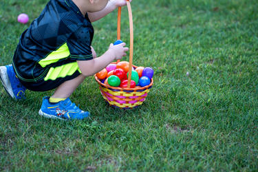 A young child crouches on green grass, placing colorful plastic Easter eggs into a woven basket during an outdoor egg hunt.